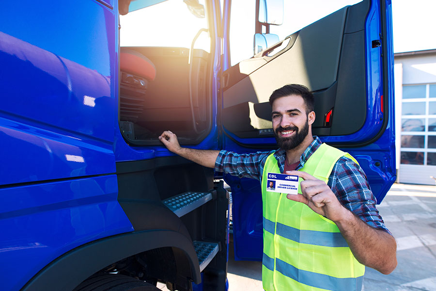 A semi-truck driver standing next to a blue commercial truck and wearing a yellow vest while holding a CDL license that was given after training.