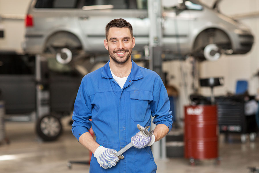 A smiling male mechanic in a blue uniform holding a wrench with a car behind him in Decatur, IL.