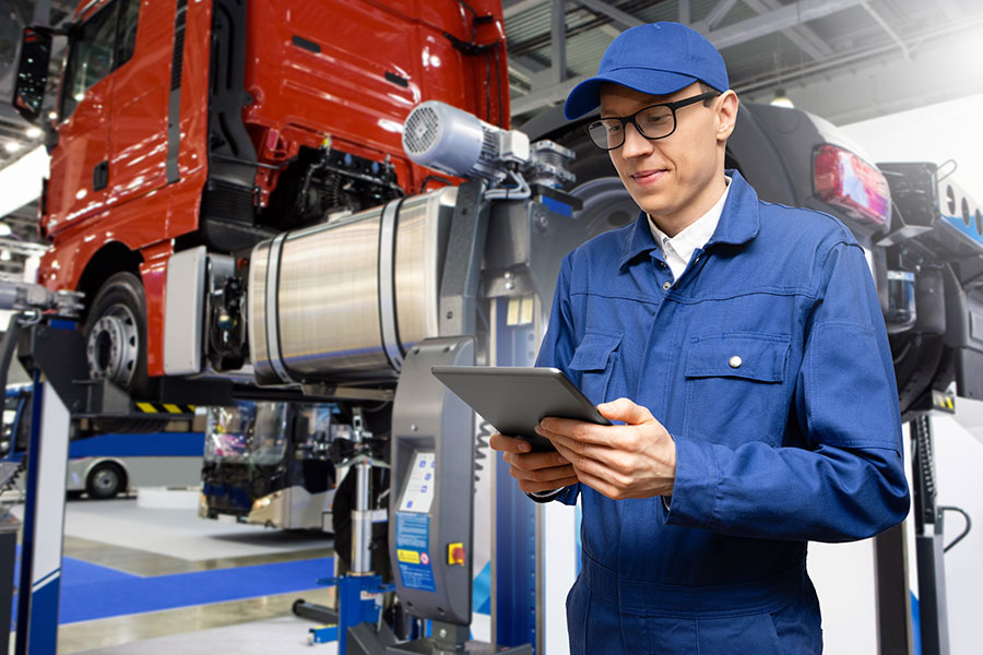 A qualified male technician in a blue uniform examining the electric system of a semi-truck and making notes on an iPad about the electrical diagnostics in Decatur, IL.