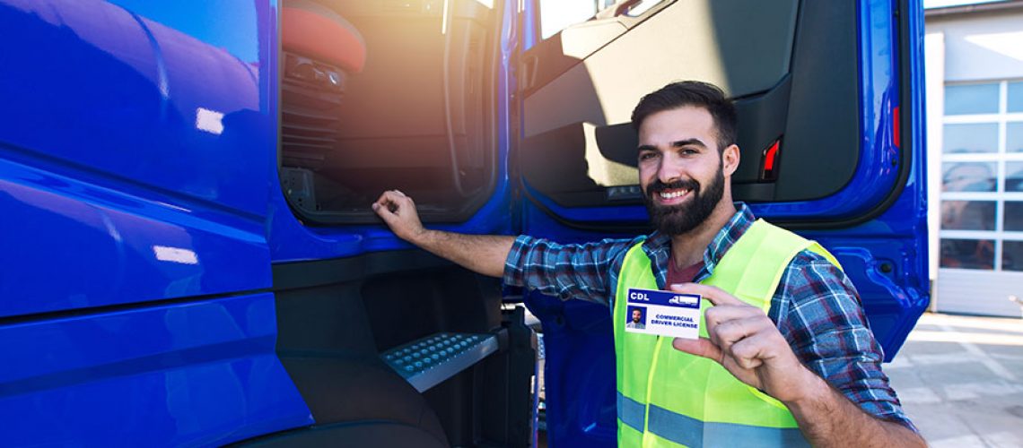 A semi-truck driver standing next to a blue commercial truck and wearing a yellow vest while holding a CDL license that was given after training.